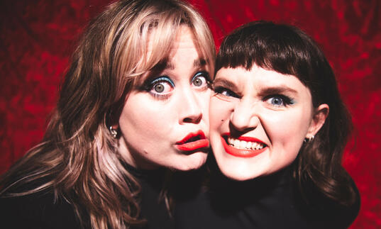 Two people wearing black against a red background, smiling with cheeks pressed together
