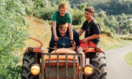Three teenagers ride on top of a red tractor, grinning