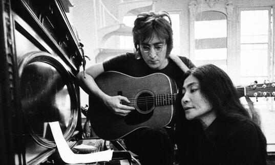 John Lennon and Yoko Ono sit at a piano, reading music in their New York apartment