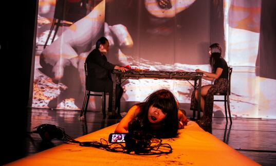 Woman on orange-lit stage with camera; large projected face creates surreal, haunting atmosphere.