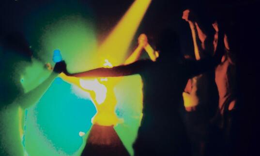 Figures are dancing in a dark room lit by yellow, green and blue lights.