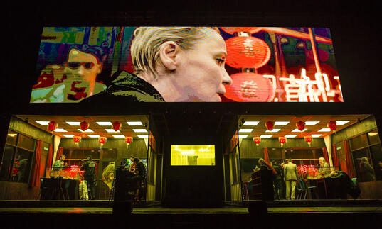 People move around in a chinese restaurant as a close-up image of one person in the venue is projected on a large screen above the set.