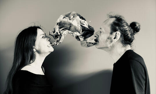 Two people stand opposite each other, a material shape - part balloon, part speech bubble - hangs between them.