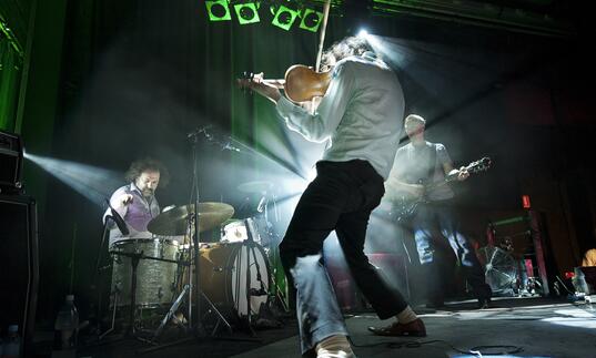 Image of a band perfoming with the guitarist's back to the camera and drummer in the background