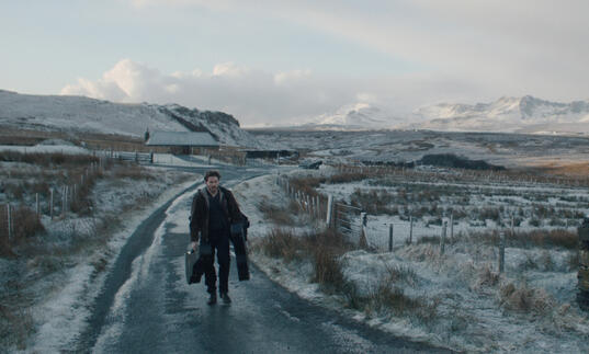 A man walks down a snowy path, holding lots of luggage