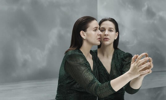 A woman on a grey stage leans against a mirror, her face reflects back at her. 