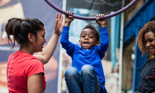 A child hanging from an aerial hoop with support from two workshop facilitators