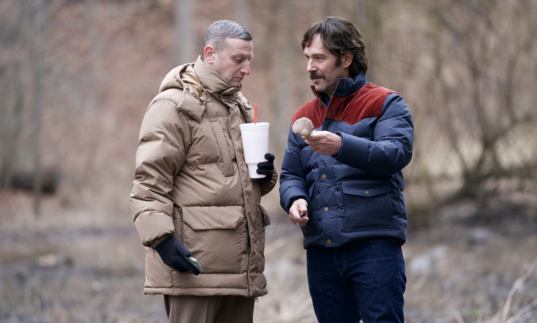 Two men in big winter coats share snacks on a walk