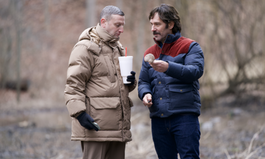 Two men in big winter coats share snacks on a walk