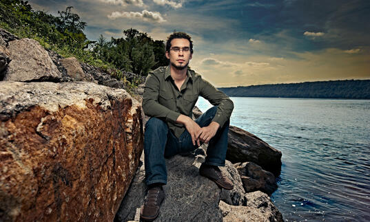 Jazz pianist and composer Fabian Almazan sits on large rocks by a serene lakeside, wearing a green shirt and jeans, with trees and a dramatic sky in the background.