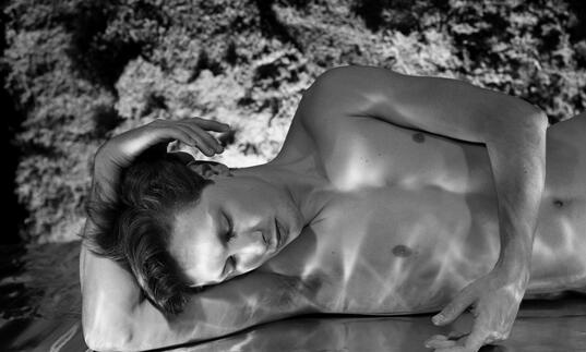A topless white man lays on a mirror covered in water, staring at his reflection