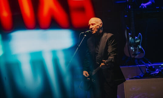 a photo of Midge Ure in a suit playing the guitar under the haze of a red and blue neon sign.
