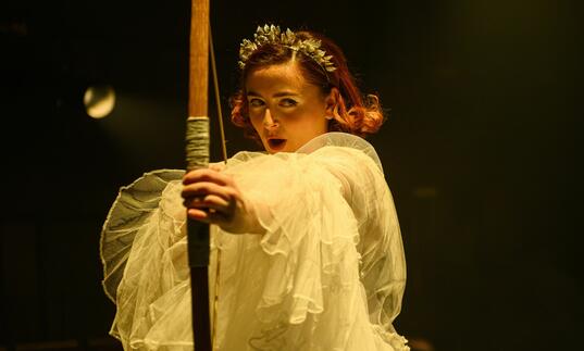 Opera singer dressed in white ruffled outfit and roman crown holding bow and arrow