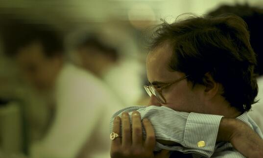 Man in smart clothing in glasses and with gold ring looks worried, covering his mouth with his arm