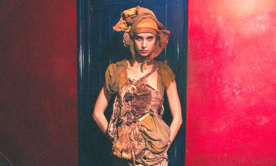 A model stands in front of a blue door and red wall, wearing earth toned distressed clothing
