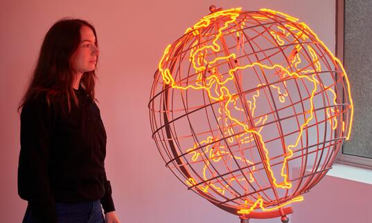 A woman looks at a sculpture of a globe, the outlines of the countries lit in neon orange strip lights.