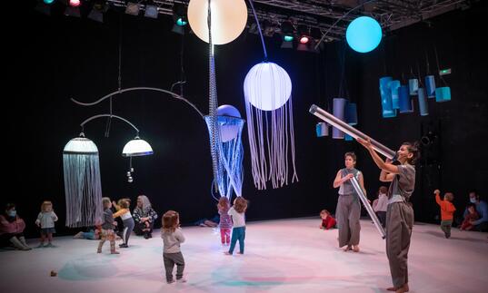 Toddlers running around with orbs of light hanging from the ceiling while two people overlook them