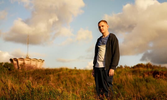 A young white man stands in a field looking into the sunset