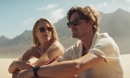 A man and a woman sit on the beach looking at each other with mountains in the background
