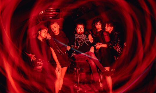 The creative team of Transpose pose alongside each other against an iconic Barbican backdrop with a red swirling light around them.