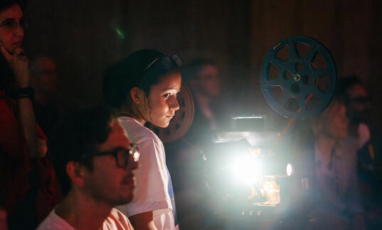 A group of people and a young child look on. A film projector emits light