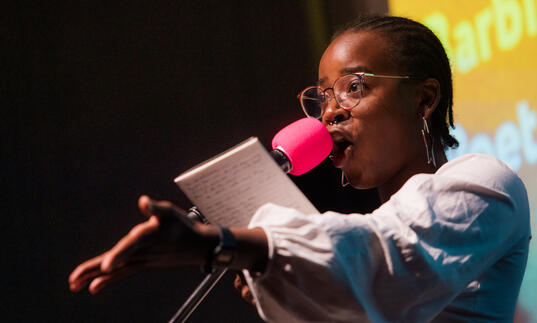 A young Black woman performing on stage speaking into a pink microphone, with her arm extended as she holds a book or paper.