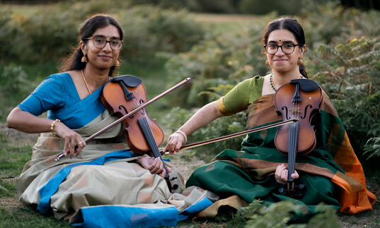 A photo of sibling duo Mathangi & Sambavi Uthayakumar sitting with violins on the grass.