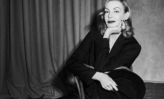 Black and white image of Ute Lemper sat backwards on a chair with her hand on her chin