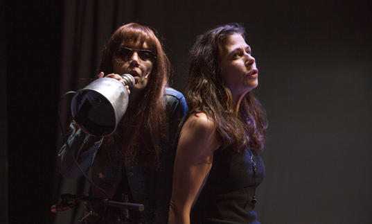 Image shows two women with long hair leaning against each other and singing