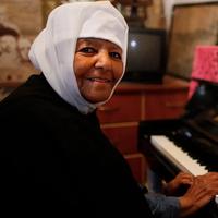 Photo of Emahoy Tsegué-Maryam Guèbrou smiling at the camera while sitting at a piano