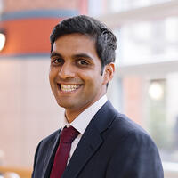 A headshot of the board member Aaron D&#039;Souza, smiling and wearing a suit and tie