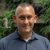 A headshot of Beau Vigushin, in a blue shirt standing in the Conservatory